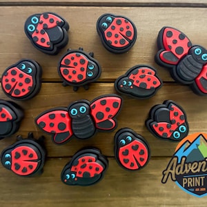 Puede incluir: Una colección de dijes para zapatos con temática de mariquitas rojas y negras. Los dijes presentan una variedad de diseños de mariquitas, incluidos algunos con alas abiertas. Las mariquitas tienen puntos negros y ojos azules.