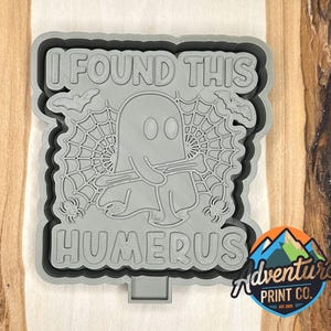 May include: Gray Halloween-themed cookie cutter with the text "I FOUND THIS HUMERUS". The design features a ghost holding a bone, spiderwebs, bats, and spiders. The Adventure Print Co. logo is in the bottom right corner.