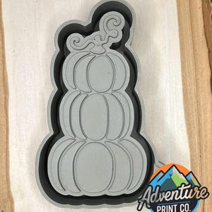Può includere: Un tagliabiscotti a forma di zucca grigia con un contorno nero. Il tagliabiscotti è progettato con tre zucche impilate e un gambo riccio. Il logo Adventure Print Co. si trova nell'angolo in basso a destra.