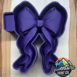 May include: A purple bow-shaped cookie cutter with a rectangular handle. The bow has detailed ribbon folds and curves. The Adventure Print Co. logo is visible in the bottom right corner.