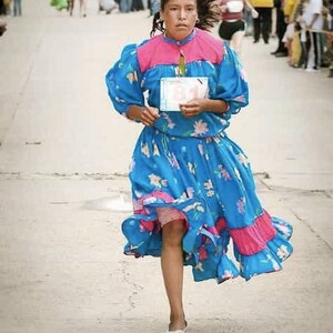 Puede incluir: Una persona joven que lleva un vestido azul y rosa con estampados florales y una etiqueta de n&uacute;mero blanca. El vestido tiene un dobladillo con volantes y la persona est&aacute; corriendo.