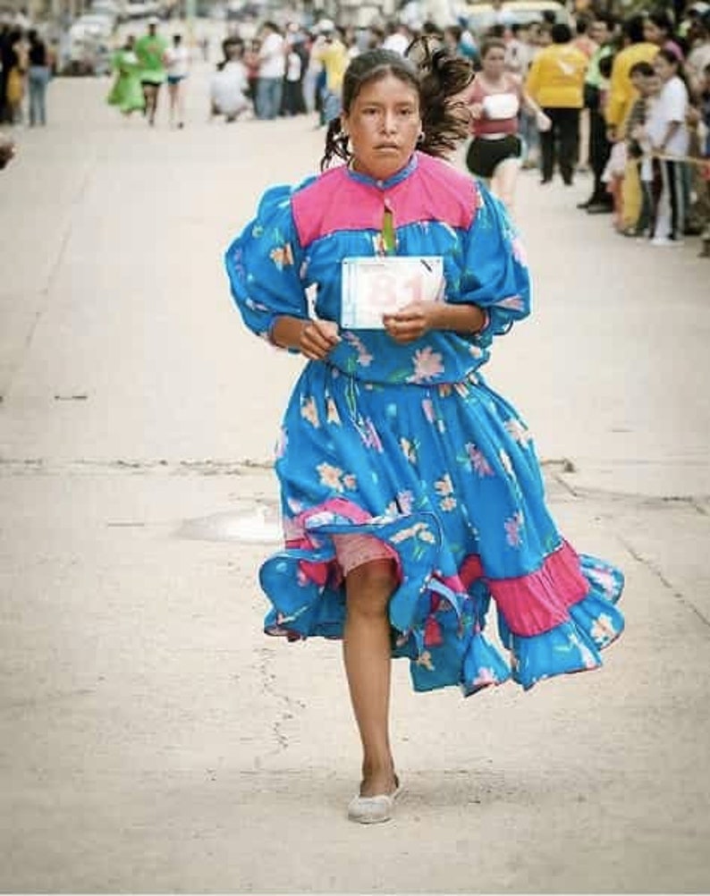 Puede incluir: Una persona joven que lleva un vestido azul y rosa con estampados florales y una etiqueta de n&uacute;mero blanca. El vestido tiene un dobladillo con volantes y la persona est&aacute; corriendo.