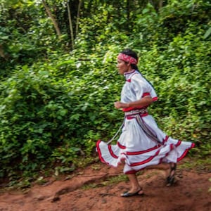 Puede incluir: Una persona vestida con un atuendo tradicional blanco con detalles rojos y negros corre por un bosque verde. El atuendo tiene una falda larga con rayas rojas y blancas y una faja roja. La persona lleva una cinta roja en la cabeza y sandalias.