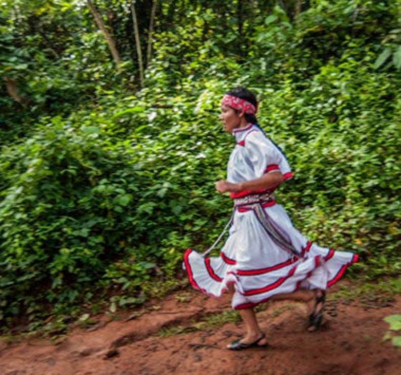 Puede incluir: Una persona vestida con un atuendo tradicional blanco con detalles rojos y negros corre por un bosque verde. El atuendo tiene una falda larga con rayas rojas y blancas y una faja roja. La persona lleva una cinta roja en la cabeza y sandalias.