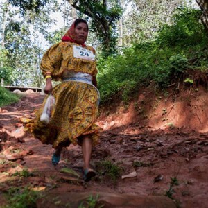 Puede incluir: Una mujer con un vestido amarillo y rojo con una faja blanca y el n&uacute;mero 24 en la parte delantera. Est&aacute; caminando por un sendero de tierra en un bosque.