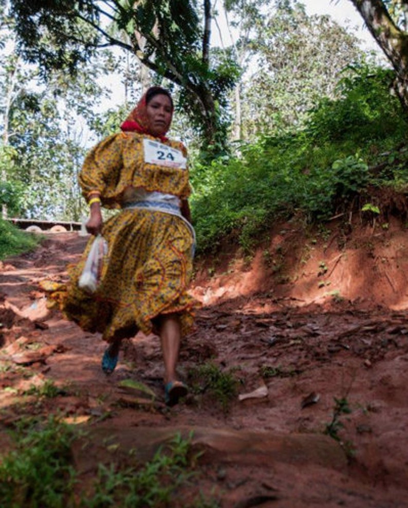Puede incluir: Una mujer con un vestido amarillo y rojo con una faja blanca y el n&uacute;mero 24 en la parte delantera. Est&aacute; caminando por un sendero de tierra en un bosque.