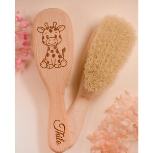Könnte beinhalten: Zwei Babybürsten aus Holz. Eine Bürste zeigt eine Giraffenillustration und den Namen "Thila" auf dem Griff. Die andere Bürste hat weiche, hellfarbene Borsten. Rosa Blumen sind im Hintergrund.