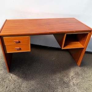 May include: A mid-century modern desk made of wood, featuring a warm reddish-brown finish. The desk has two drawers on the left and an open storage space on the right. The desk's design includes angled legs and a rectangular tabletop.