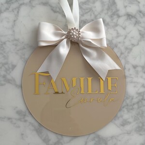 May include: A round, beige acrylic sign with gold lettering that reads "FAMILIE Emrula." A white satin ribbon and bow with a pearl and rhinestone embellishment are attached to the top for hanging.