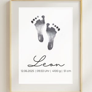 May include: Framed print featuring two grayscale baby footprints on a white background. The name "Leon" is written in cursive below the footprints, along with the date, time, weight (4100 g), and height (51 cm). The frame is light wood.