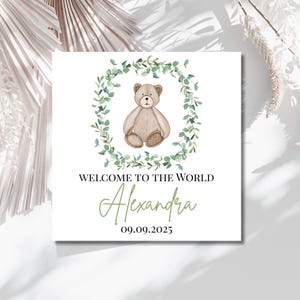 May include: A square card featuring a teddy bear illustration within a green foliage wreath. The text reads "WELCOME TO THE WORLD", followed by the name "Alexandra" and the date "09.09.2025". The design is set against a white backdrop.