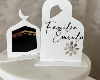 Op de afbeelding: Drie witte lasergesneden decoraties met moskee- en maansikkelontwerpen. Eén toont de Kaaba, een andere heeft de tekst "Familie Emrula" en een uitsparing in de vorm van een ster. De decoraties staan op een witte marmeren taartstandaard.