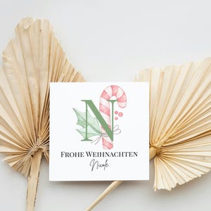 Könnte beinhalten: Eine quadratische Weihnachtskarte mit einem Aquarelldesign. Sie zeigt den grünen Buchstaben „N“, eine Zuckerstange und Stechpalmenblätter. Die Karte trägt den Text „FROHE WEIHNACHTEN Nicole“ in schwarzer Schrift. Die Karte ist vor getrockneten Palmblättern platziert.