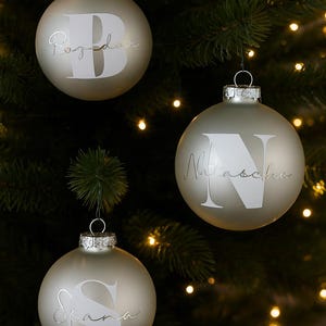 Könnte beinhalten: Drei Weihnachtsbaumkugeln aus mattiertem Glas, die an einem Baum hängen. Jede Kugel hat einen silberfarbenen Buchstaben und Namen: "Bozidar", "Nattascha" und "Siana". Die Kugeln sind an silbernen Metallkappen aufgehängt.