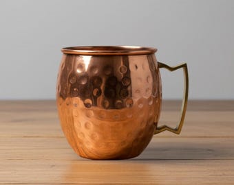 May include: A copper Moscow Mule mug with a hammered finish and a brass handle. The mug sits on a wooden surface. The mug is designed for serving cold beverages.