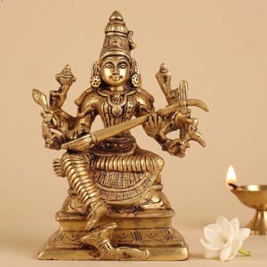 May include: A detailed brass statue of a seated deity, adorned with intricate jewelry and holding a stringed instrument. The statue has multiple arms and is set on a decorative base. A small oil lamp and a white flower are in the background.