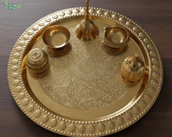 May include: A gold-colored brass puja thali set. The set includes a round tray with a decorative border, small bowls, a bell, and a container. The tray has an embossed floral pattern. The set is designed for religious ceremonies.