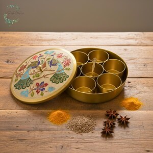 Puede incluir: Una caja de especias dorada con una tapa decorada con pavos reales y dise&ntilde;os florales. La caja contiene varios compartimentos peque&ntilde;os y una cuchara. Especias como c&uacute;rcuma, comino y an&iacute;s estrellado est&aacute;n dispuestas sobre una superficie de madera.