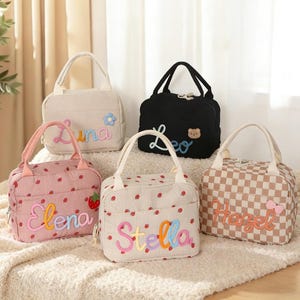 Pode incluir: Cinco lancheiras de veludo cotelê em várias cores. Uma é preta, uma é rosa com detalhes de morango, uma é bege com detalhes de morango, uma é bege com uma flor e uma é xadrez. Cada saco tem um nome bordado.