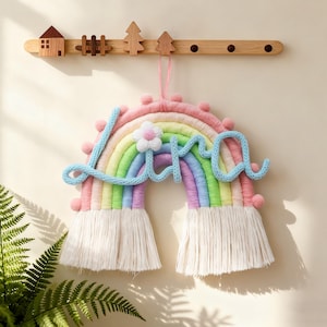 Puede incluir: Un adorno de pared de arcoíris hecho a mano con el nombre "Lara" en hilo azul claro. El arcoíris presenta colores pastel, una flor blanca y flecos en la parte inferior. Elementos decorativos de madera, como una casa, una valla y árboles, están arriba.