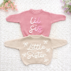 Può includere: Due maglioni per bambini. Il maglione superiore è rosa con le parole "Lil Sis" in corsivo bianco. Il maglione inferiore è beige con le parole "Little Sister" in corsivo bianco e dettagli di fiocchi bianchi. Entrambi i maglioni hanno uno scollo tondo.