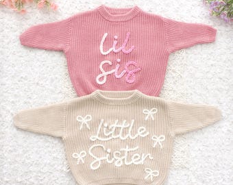 May include: Two children's sweaters. The top sweater is pink with the words "Lil Sis" in white script. The bottom sweater is tan with the words "Little Sister" in white script and white bow accents. Both sweaters have a round neckline.
