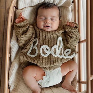 May include: A baby wearing a long-sleeved, olive-green knitted jumper with the name Bodie embroidered in white cursive yarn across the chest. The jumper features a ribbed texture and is worn over a white nappy while the baby sleeps.