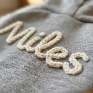 Może przedstawiać: Zbliżenie szarej bluzy z kapturem z imieniem "Miles" wyhaftowanym kremową, teksturowaną przędzą. Litery są w stylu kursywy, dodając spersonalizowany akcent do ubrania. Materiał bluzy wydaje się miękki i wygodny.