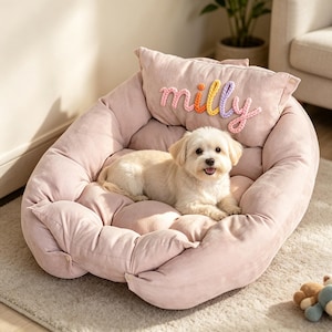 Puede incluir: Una cama para mascotas de color rosa claro, acolchada y de forma ovalada, con bordes elevados y un cojín rectangular a juego en la parte posterior. El cojín presenta el nombre "milly" bordado con letras de hilo trenzado y colorido.
