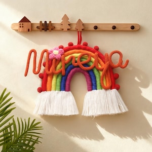 May include: A vibrant, rainbow-shaped wall hanging spelling "Nireya" in orange yarn, with a pink flower detail. The rainbow displays a spectrum of colours and white fringe. Above, a wooden wall hook with decorative wooden shapes.