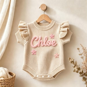Könnte beinhalten: Cremefarbener gestrickter Baby-Strampler mit dem Namen "Chloe" in rosa Schrift. Der Strampler hat Rüschenärmel und drei rosa Blumenapplikationen. Er hängt an einem Holzbügel vor einem neutralen Hintergrund.