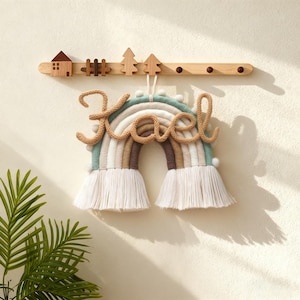 Puede incluir: Un adorno de pared de arcoíris hecho a mano con el nombre "Kael" en cursiva. El arcoíris presenta colores pastel y una franja en la parte inferior. Encima, un colgador de pared de madera con una pequeña casa, valla y árboles.