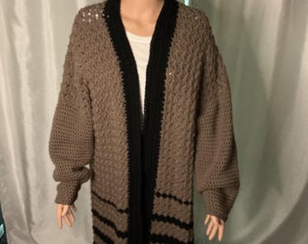 May include: A long, handmade, brown crochet cardigan with black trim and stripes. The open-front design features a textured stitch pattern and long sleeves. The cardigan is displayed on a mannequin.