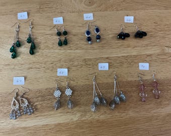 Puede incluir: Una colección de varios pendientes exhibidos sobre una superficie de madera. Los pendientes presentan diversos diseños, incluyendo cuentas colgantes, formas florales y trabajos de metal intrincados. Los colores van desde el verde esmeralda y el negro hasta el plateado y el rosa.