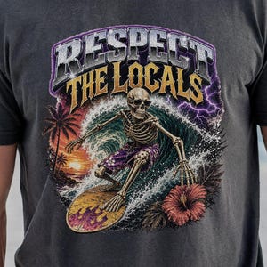 May include: A charcoal gray t-shirt featuring a graphic print of a skeleton surfing on a wave. The text "RESPECT THE LOCALS" appears in bold lettering above the skeleton. The design includes a sunset, palm trees, and a red hibiscus flower.