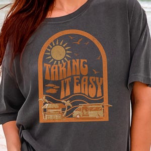 May include: Dark gray t-shirt with a retro design. The design features a sun, palm trees, and the words "TAKING IT EASY" in orange. The shirt is a relaxed fit, perfect for a casual day at the beach or a road trip.