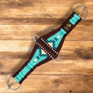 May include: A turquoise, brown, and cream-colored horse cinch with metal rings. The cinch features a geometric pattern in the center and is branded with the text "IQRO UNIQUE" and a horse logo on a brown leather patch.