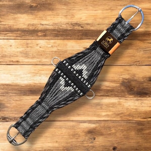 May include: A black and gray woven horse cinch with silver-colored buckles and rings. The cinch features a geometric pattern and a leather patch with the text "IQRO UNIQUE" and a horse silhouette.