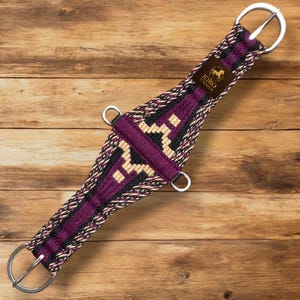 Puede incluir: Una cincha de caballo morada, negra y beige con herrajes de metal plateado. La cincha presenta un diseño tejido con un patrón geométrico y el texto "JORO UNIQUE" en una etiqueta marrón.