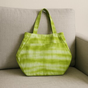 May include: A lime green tote bag with a horizontal tie-dye pattern. The bag features a hexagonal shape with a wide base, two matching fabric handles, and a large exterior slip pocket across the front panel, constructed from soft textile.
