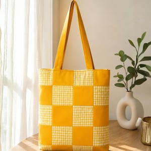 May include: A tote bag featuring a checkerboard pattern of solid yellow squares and white and yellow gingham squares. The bag has two long yellow shoulder straps and a rectangular body constructed from alternating fabric blocks in a grid layout.