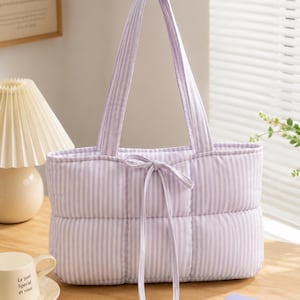 May include: A quilted tote bag featuring a light purple and white vertical striped pattern. The bag has two shoulder straps and a central drawstring closure with two long fabric ties hanging down the front. The rectangular body is padded.