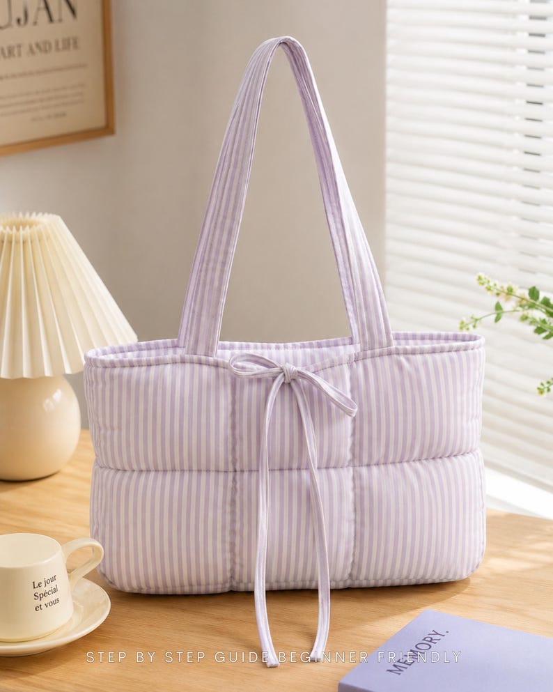 May include: A quilted tote bag featuring a light purple and white vertical striped pattern. The bag has two shoulder straps and a central drawstring closure with two long fabric ties hanging down the front. The rectangular body is padded.