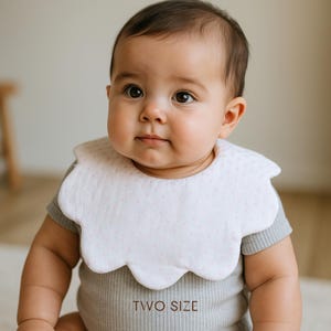 Puede incluir: Un bebé con un babero blanco con borde festoneado y pequeños lunares rosas. El babero está sobre un body gris acanalado. El texto "TWO SIZE" está impreso en el body.