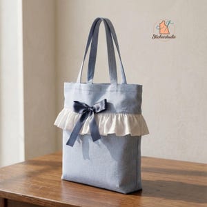 Può includere: Una borsa tote in tessuto azzurro con un bordo a volant bianco sul davanti e un nastro di raso blu scuro annodato a fiocco al centro. La borsa è dotata di due lunghi manici a spalla e include un piccolo logo con la scritta Stichestudio.