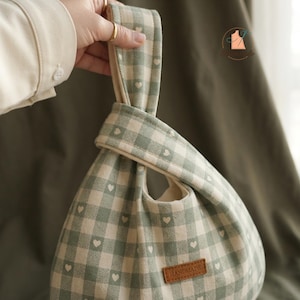 May include: A sage green and off-white gingham fabric bag featuring a small heart pattern throughout. The bag has a single continuous handle loop design and a rectangular brown leather-like tag on the front with the word HANDMADE embossed.