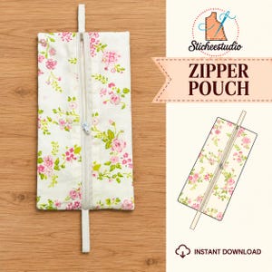 May include: A rectangular zipper pouch featuring a white fabric base with a pink and green floral pattern. The pouch includes a central vertical zipper with a white pull and a white fabric tab at the top and bottom. Text reads Sticheestudio ZIPPER POUCH.