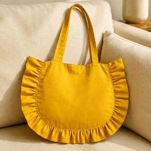 Puede incluir: Un bolso tote de color amarillo mostaza con una base redondeada y un borde de tela con volantes en los laterales. El bolso incluye dos asas de tela a juego y el texto STEP BY STEP GUIDE BEGINNER FRIENDLY en la parte inferior.