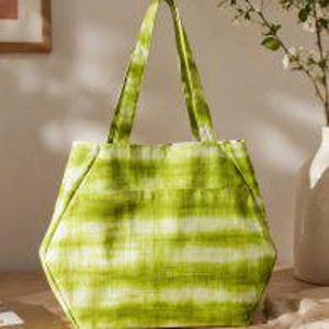 May include: A lime green tote bag featuring a horizontal tie-dye or watercolor-style stripe pattern. The bag has a structured, hexagonal shape with two integrated fabric handles at the top and a flat base, constructed from a soft textile material.