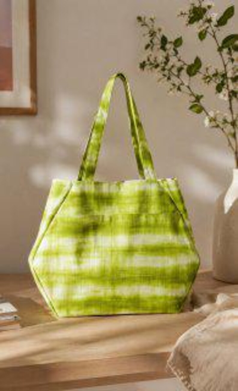 May include: A lime green tote bag featuring a horizontal tie-dye or watercolor-style stripe pattern. The bag has a structured, hexagonal shape with two integrated fabric handles at the top and a flat base, constructed from a soft textile material.